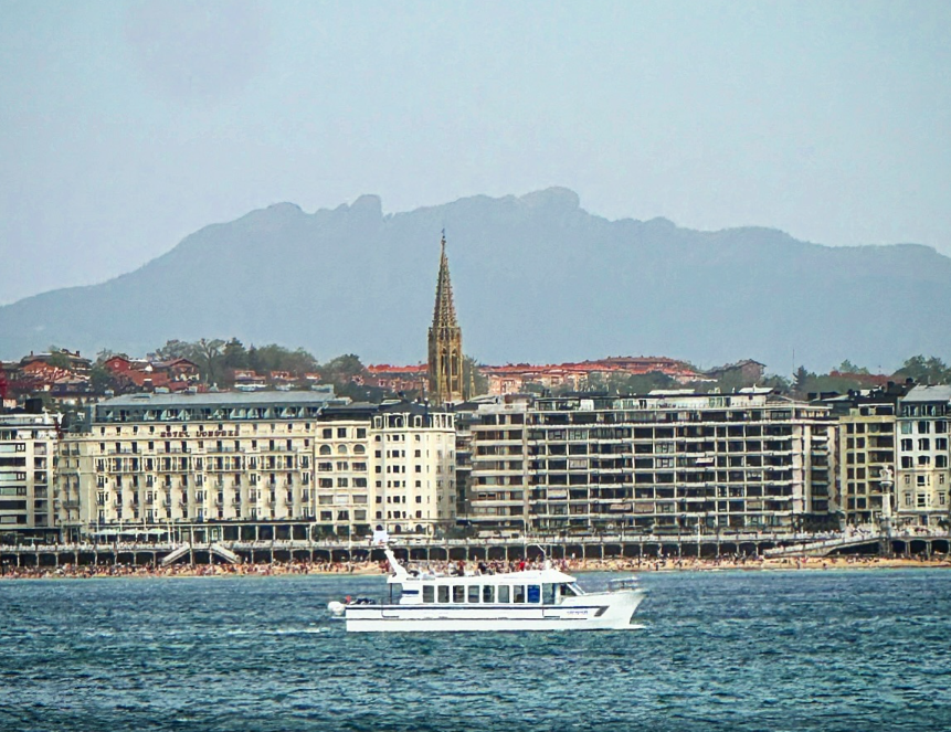 Vista de la Bahía de La Concha desde el mar con catamarán turístico en primer plano, edificios Belle Époque, aguja de la Catedral del Buen Pastor y monte Urgull al fondo